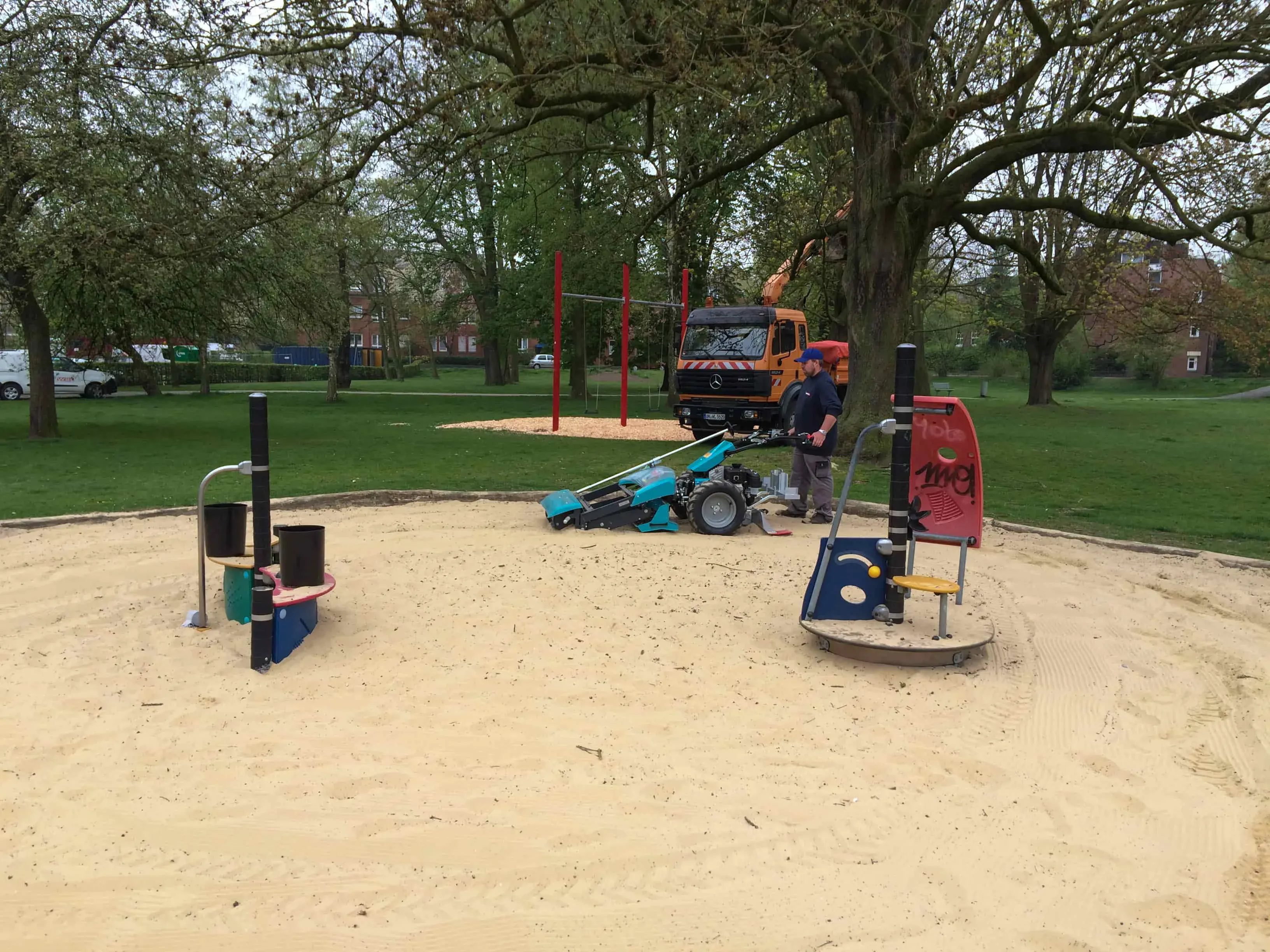 Mann reinigt einen Spielplatzsandbereich mit einer BeachTech Maschine, im Hintergrund Bäume und Parkanlage.
