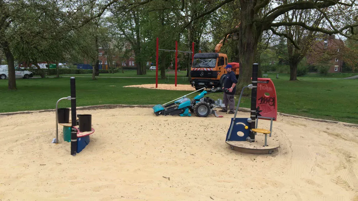 BeachTech Maschine reinigt einen Spielplatzsandbereich; im Vordergrund Spielgerät, im Hintergrund Park und Bäume.