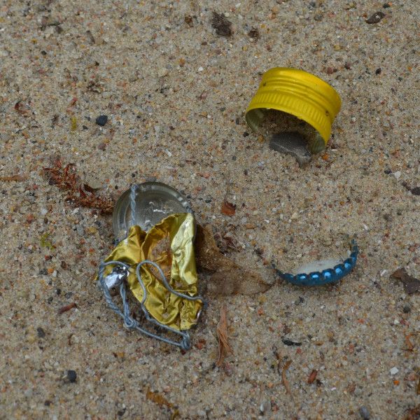 Mehrere Kronkorken und zerknüllte Metallteile liegen verstreut im Sand.