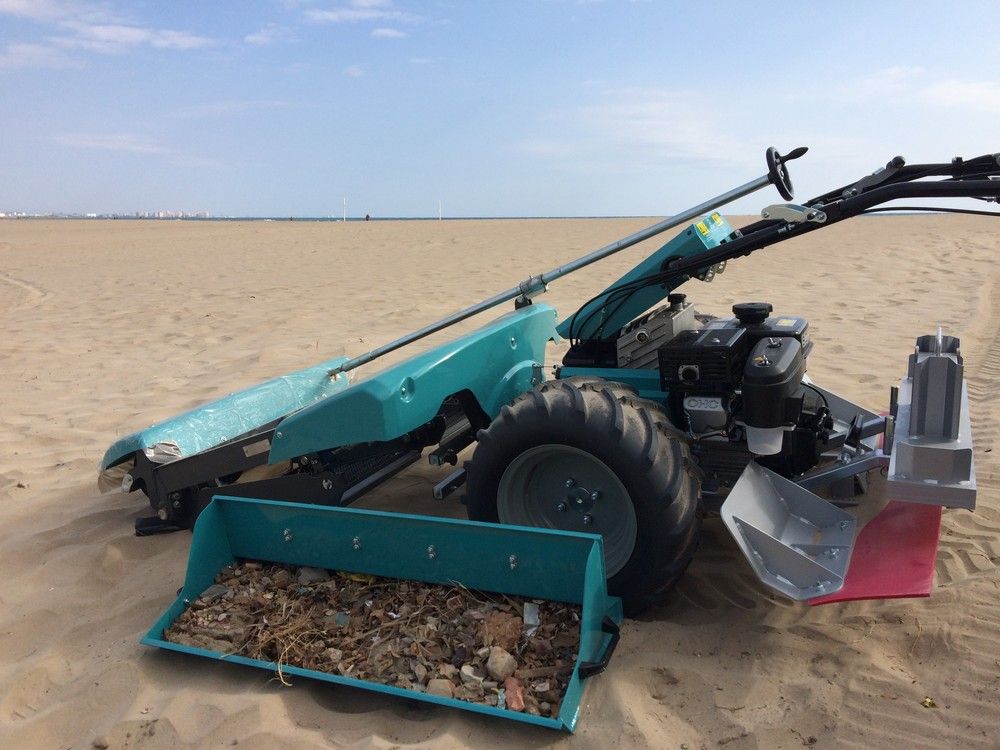 BeachTech Sweepy auf einem Sandstrand, die Müll und Ablagerungen in einem Auffangbehälter gesammelt hat. Im Hintergrund ist das Meer und ein klarer Himmel zu sehen.