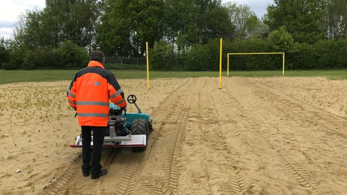 Mann in orangefarbener Warnjacke bedient eine BeachTech-Reinigungsmaschine auf einem sandigen Sportfeld mit gelben Torpfosten im Hintergrund