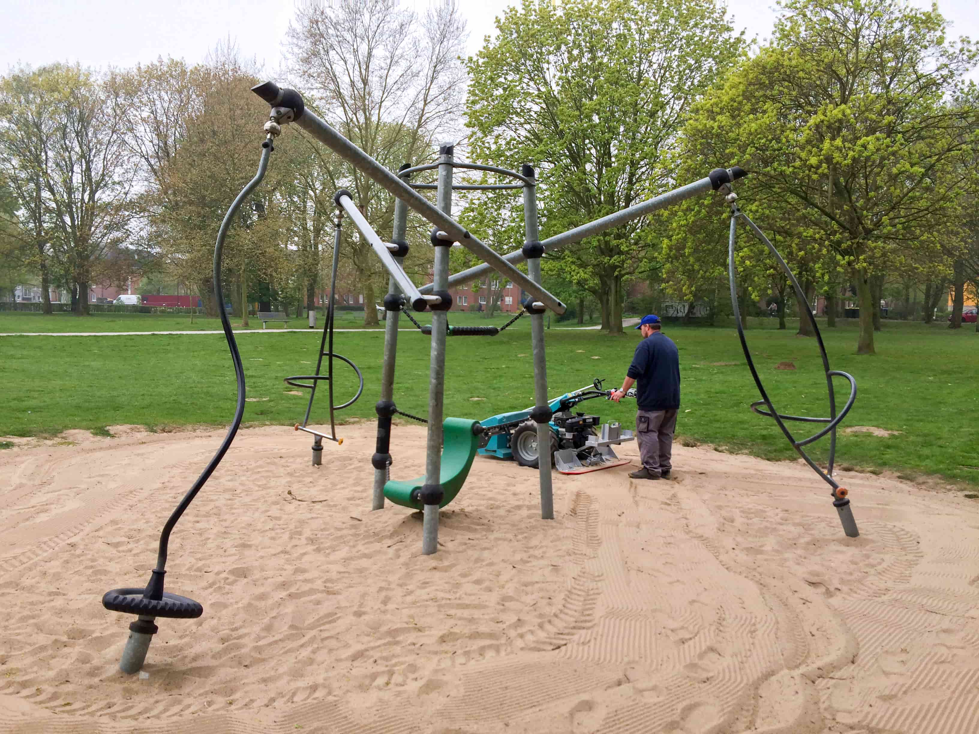 Mann reinigt einen Sandspielbereich mit BeachTech Sweepy Hydro, im Vordergrund Klettergerüst auf sauberem Sand.